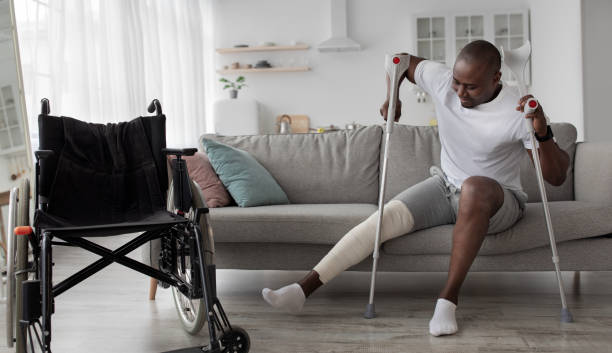 Patient is recuperating in living room due to illness. Concentrated cheerful tense adult african american guy with broken leg in cast stands up with crutches from couch near wheelchair, panorama