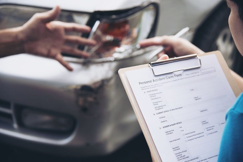 A person holds a clipboard with a car accident form near a damaged vehicle.